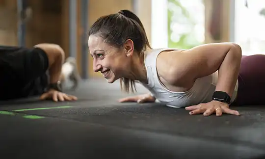 A smiling woman performs push-ups during a group fitness workout, building upper-body strength and endurance at Krav Maga Chapel Hill.